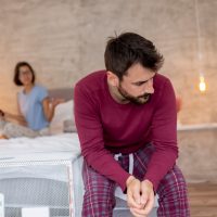 Couple fighting in bedroom, woman using a smartphone in the background while man sitting in the foreground, sad and pensive