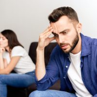 Upset young bearded man in blue shirt sitting on side opposite to girlfriend and rubbing forehead while suffering about fight